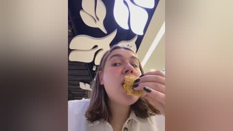 Media: A video of a young woman with light skin and shoulder-length brown hair, wearing a white shirt, eating a honey-covered biscuit in a brightly lit room with a black and white abstract mural in the background.