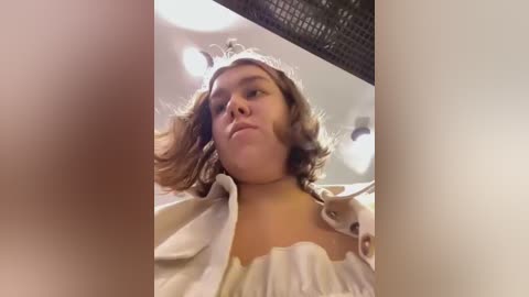 Media: Video of a young woman with shoulder-length, wavy brown hair, wearing a beige top, looking up from a low angle, with blurred background and ceiling lights.