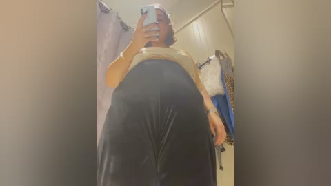 Media: Video of a plus-sized woman with short brown hair, wearing a beige top and black shiny leggings, taking a selfie in a dimly lit room with a white curtain and hanging clothes.