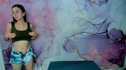 Media: Video of a young woman with straight brown hair, wearing a green crop top and blue patterned shorts, standing in a room with a vibrant, abstract, pastel-colored mural on the wall.