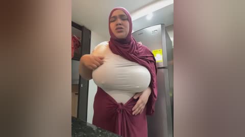 Media: A video of a plus-sized woman with large breasts, wearing a burgundy hijab and matching dress, adjusting her top in a modern kitchen with stainless steel appliances and dark countertops.