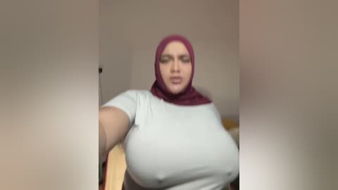 Media: Video of a plus-sized woman with large breasts, wearing a tight white t-shirt and a maroon hijab, posing indoors with a blurred background.