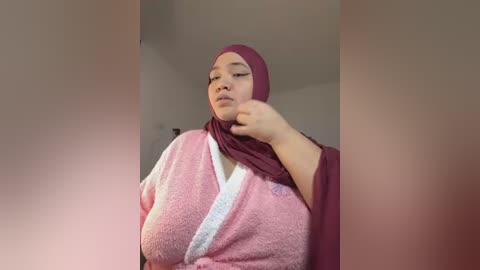 Media: A video shows a plus-sized woman with light brown skin and a maroon hijab. She wears a pink bathrobe and adjusts her hijab with a hand. The background is a simple, beige indoor setting.