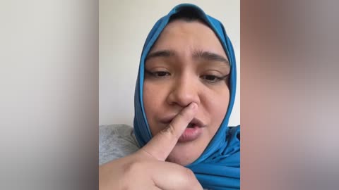 Media: Video of a young woman with medium skin tone, dark hair, and brown eyes, wearing a blue hijab. She is playfully covering her mouth with her index finger, set against a plain, light-colored background.