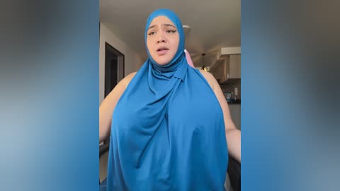 Media: Video of a plus-sized woman with a light brown complexion, wearing a bright blue hijab and headscarf, indoors with blurred background showing a kitchen and living area.