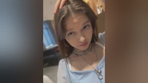 Media: A video of a young woman with light skin and long brown hair, wearing a light blue top and silver necklace, touching her head, in a dimly lit room.