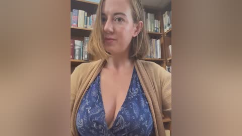 Media: Video of a light-skinned woman with shoulder-length blonde hair, wearing a low-cut blue top with white patterns, and a beige cardigan, in a library with wooden bookshelves filled with various books in the background.