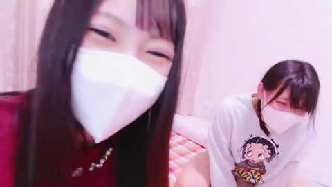 Video of two women wearing white face masks, one with long black hair, the other with short dark hair. They are indoors with a soft pink background.