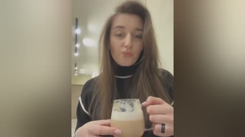 Media: Video of a young woman with long brown hair, wearing a black turtleneck, blowing a kiss while holding a glass of beer with a foamy head. Background features a blurred bathroom with a mirror and wall-mounted lights.