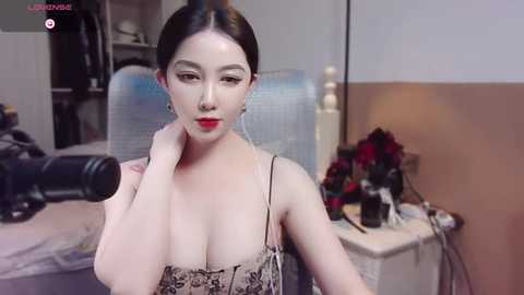 Media: A video of a fair-skinned Asian woman with long black hair, wearing a low-cut black and white floral dress, posing in a bedroom with a camera, a desk, and a bed in the background.