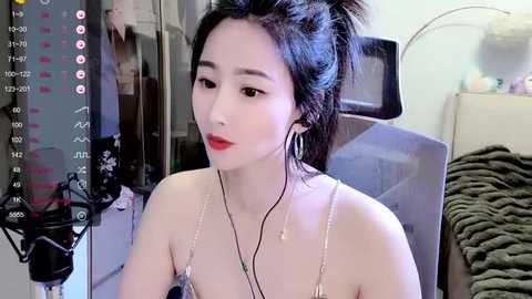 Media: Video of an Asian woman with fair skin and dark hair in a messy bun, wearing red lipstick and a spaghetti strap top, seated indoors, looking at a phone.