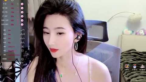 Media: Video of an Asian woman with long black hair, fair skin, and red lipstick, wearing a sleeveless top, sitting in a chair with headphones. Background shows a cluttered room with a stuffed animal and a messy desk.