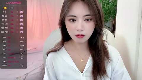 Media: A video of a young Asian woman with long, straight brown hair, fair skin, and red lipstick, wearing a white shirt, in a bedroom with a green plant.