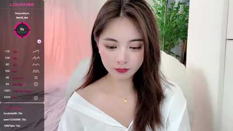 Media: Video of a young Asian woman with fair skin, long brown hair, and an off-shoulder white top, seated indoors with a green plant behind her.