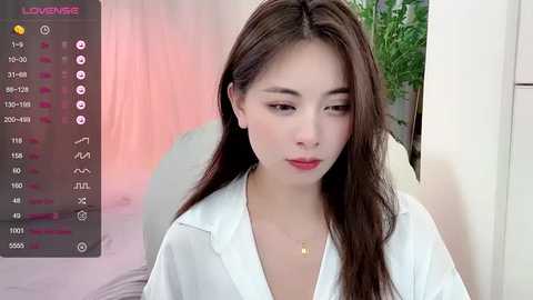 Media: A video of a young Asian woman with long dark hair, fair skin, and light makeup, wearing a white blouse, against a blurred indoor background with a plant.