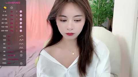 Media: A video of a young Asian woman with long brown hair and fair skin, wearing a white off-shoulder blouse, sitting indoors with green plants in the background.