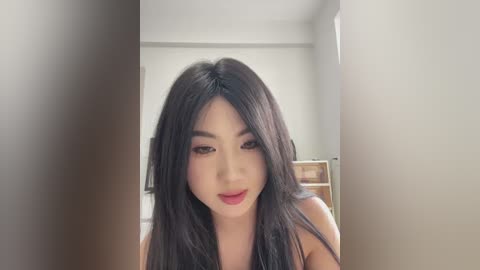 Media: Video of an Asian woman with long black hair, fair skin, and red lipstick, smiling in a minimalistic, white-walled room with a wooden shelf in the background.