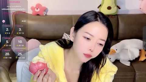 Media: A video of an East Asian woman with long black hair, wearing a yellow jacket, holding a red heart-shaped dessert. Background includes a brown couch with plush toys, including a duck and bear.