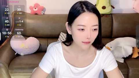 Media: Video of a young East Asian woman with straight black hair, wearing a white t-shirt, sitting on a brown leather couch with plush toys, including a bear and a duck.