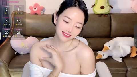 Media: A video of a smiling East Asian woman with long black hair, wearing a white off-shoulder top, sitting on a brown leather couch surrounded by stuffed animals.