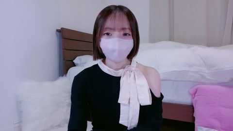 Media: Video of a young Asian woman with short brown hair, wearing a black dress with a white bow, a white face mask, and sitting on a fluffy white rug in a minimalist bedroom with a white bed and pink pillow.