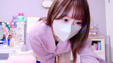 Media: Video of an Asian woman with long brown hair and bangs wearing a pink robe and white face mask, bending over a table in a tidy, pastel-toned room.