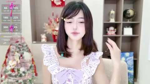 Video of a young Asian woman with straight black hair, wearing a lavender floral dress, in a modern, neatly organized living room with shelves and a decorated Christmas tree in the background.