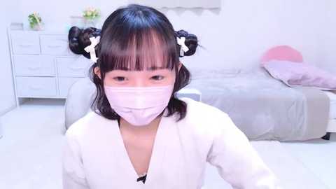 Media: A video of an Asian girl with dark hair styled in twin pigtails, wearing a pink face mask, sitting on a white chair in a minimalistic, clean bedroom with a bed and white dresser.