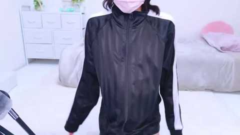 Video of a person wearing a black and white zip-up jacket in a minimalist, brightly lit room with white walls, a dresser, and a bed with light bedding.