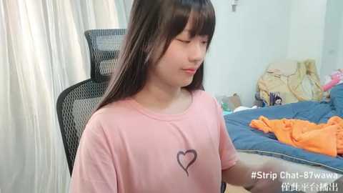 Media: Video of a young Asian woman with long dark hair, wearing a pink T-shirt with a heart graphic, sitting on a black mesh office chair in a messy bedroom.