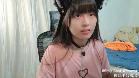 Media: A video of a young Asian woman with long dark hair, wearing a pink shirt, cat ears, and a black choker, sitting in a chair in a messy bedroom.