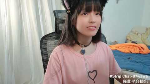 Media: Video of an Asian woman in a pastel pink T-shirt with a heart design, black cat ears, and a black choker, sitting in a black chair with a blue blanket in a bedroom.