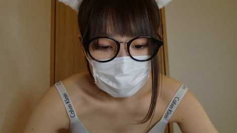 Media: Video of an East Asian woman with long black hair, wearing a white Calvin Klein bra, black-framed glasses, and a white face mask. She stands against a beige wall, looking downward.