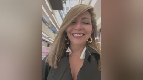 Media: Video of a smiling, fair-skinned woman with long blonde hair, wearing a black blazer, large gold hoop earrings, and a white shirt, standing in a brightly lit, modern retail setting with shelves in the background.