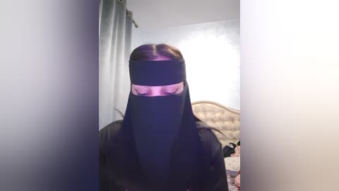 Media: Video of a person in a black niqab, standing in a dimly lit bedroom with a tufted headboard and a bed covered in a floral-patterned duvet.