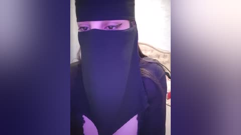 Media: Video of a woman with light skin and long brown hair wearing a black burqa with a white floral pattern, standing indoors with blurred background.