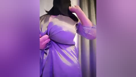 Media: A video of a woman with long black hair, wearing a shiny, light purple satin dress with a large bow at the waist. She stands in a dimly lit room with purple lighting.