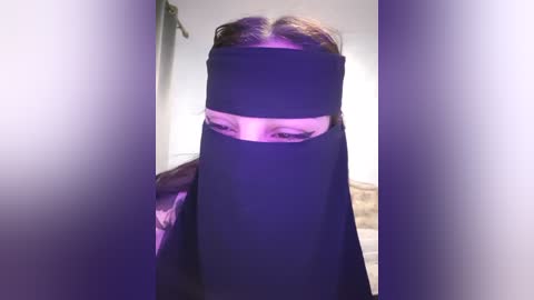 Media: A video of a person in a black niqab with a slit for the eyes, standing in a dimly lit room with purple lighting, partially visible wooden floor, and a blurred background.