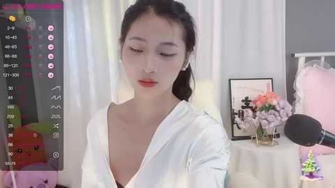 Media: Video of an Asian woman with dark hair, wearing a white blouse, recording herself in a room with a floral arrangement, pink curtains, and a microphone.