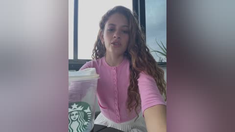 Media: Video of a young woman with curly brown hair, wearing a pink button-up top, holding a Starbucks cup, sitting in front of a window in a dimly lit room.