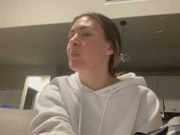 Media: Video of a woman with straight, light brown hair, wearing a white hoodie, looking serious in a modern, dimly lit kitchen with stainless steel appliances and beige walls.