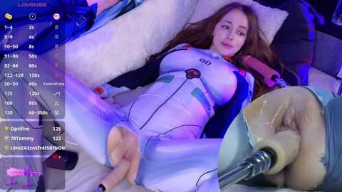 Media: Video of a woman in a white, skin-tight bodysuit, lying on a plush couch, with a dildo inserted into her vagina, surrounded by digital text and video game interface.