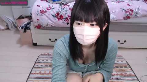 Media: A video of an Asian woman with straight black hair and a white face mask, sitting on a patterned rug in a cluttered bedroom with a floral bedspread.