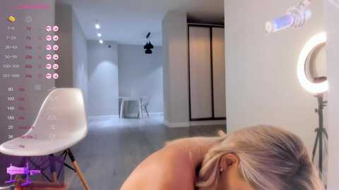 Media: Video of a nude woman with blonde hair, lying face down on a white chair in a modern, minimalist room with white walls and hardwood floors.