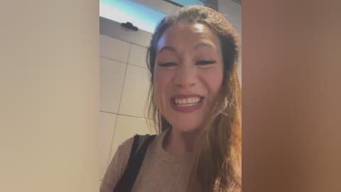 Media: Video of a smiling Asian woman with light skin, long brown hair, and a beige sweater, standing in a tiled bathroom with a window.