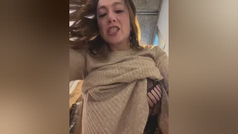 Media: Video of a young woman with fair skin and light brown hair, wearing a beige sweater and black fishnet top, playfully sticking her tongue out.