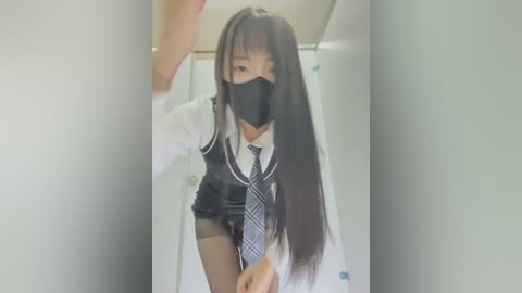 Media: Video of an Asian woman in a white lab coat, black tie, and mask, crouching in a dimly lit bathroom, looking through a gap.