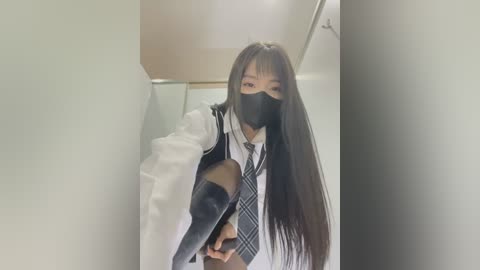 Media: Video of a young Asian woman in a white lab coat, black face mask, and plaid tie, with long black hair, standing in a narrow, bright hallway.