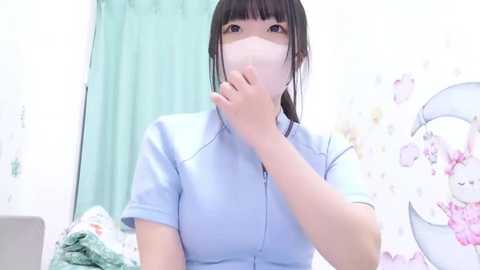 Media: Video of an East Asian woman with straight black hair and a light blue button-up shirt, wearing a white face mask. She's in a pastel-themed bedroom with a pink floral-patterned wall and light blue curtains.