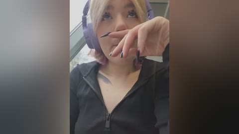 Media: Video of a young Caucasian woman with blonde hair, wearing purple headphones and a black zip-up hoodie, holding a pen to her lips, indoors.